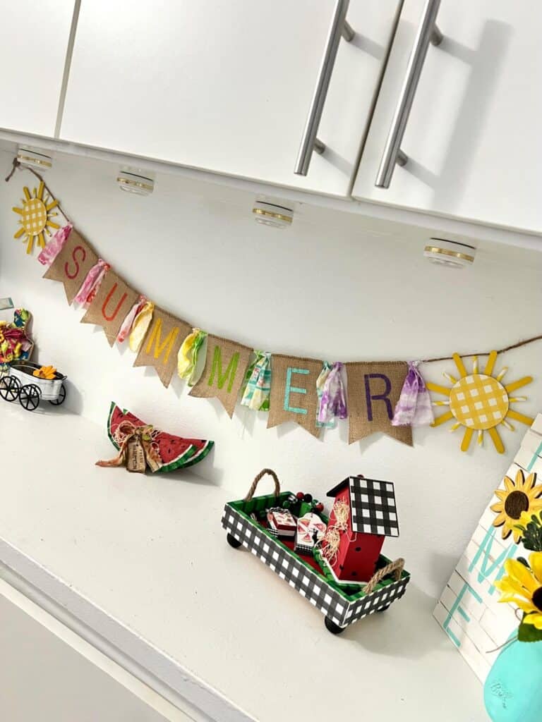 A crafty Summer burlap banner with colorful letters  stenciled on the burlap and fabric strips in between, a yellow sun on each end, hanging on a shelf decorated for summer.