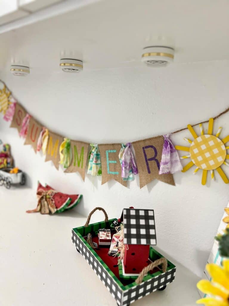 A crafty Summer burlap banner with colorful letters  stenciled on the burlap and fabric strips in between, a yellow sun on each end, hanging on a shelf decorated for summer.