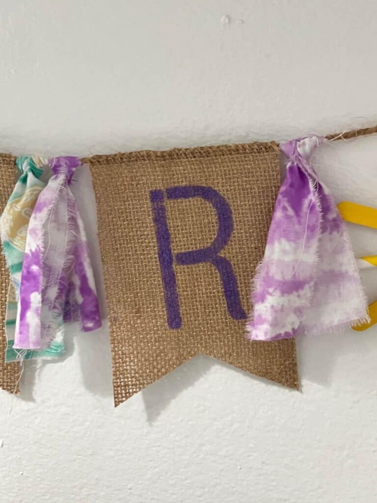 A purple letter "R" stenciled onto a burlap pendant with purple strips of fabric on each side.