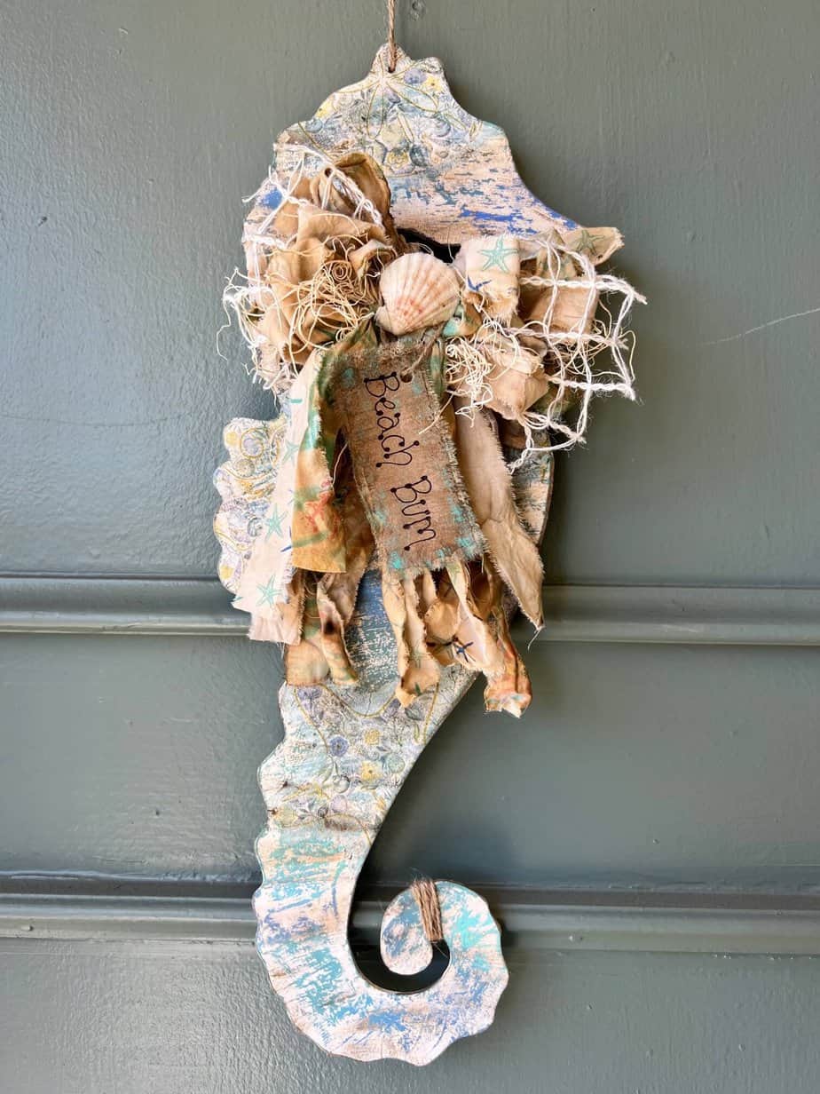 Dollar tree chippy seahorse with craftkins makeover of wood seahorse with blue green chipped paint, a seashell napkin decoupaged on it, a big messy fabric bow with a kraft paper tag that says "Beach Bum" hanging on a front door as summer coastal nautical DIY decor.