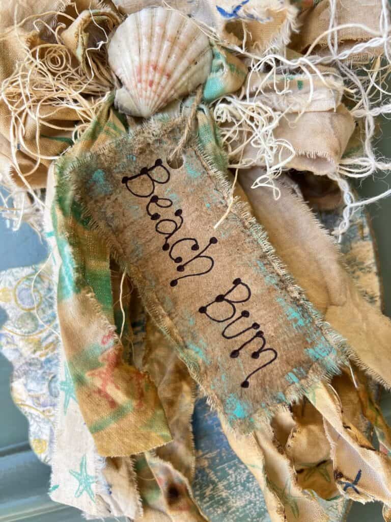 A kraft paper and muslin hangtag that says "Beach Bum" with blue and green paint distressing around the edges.