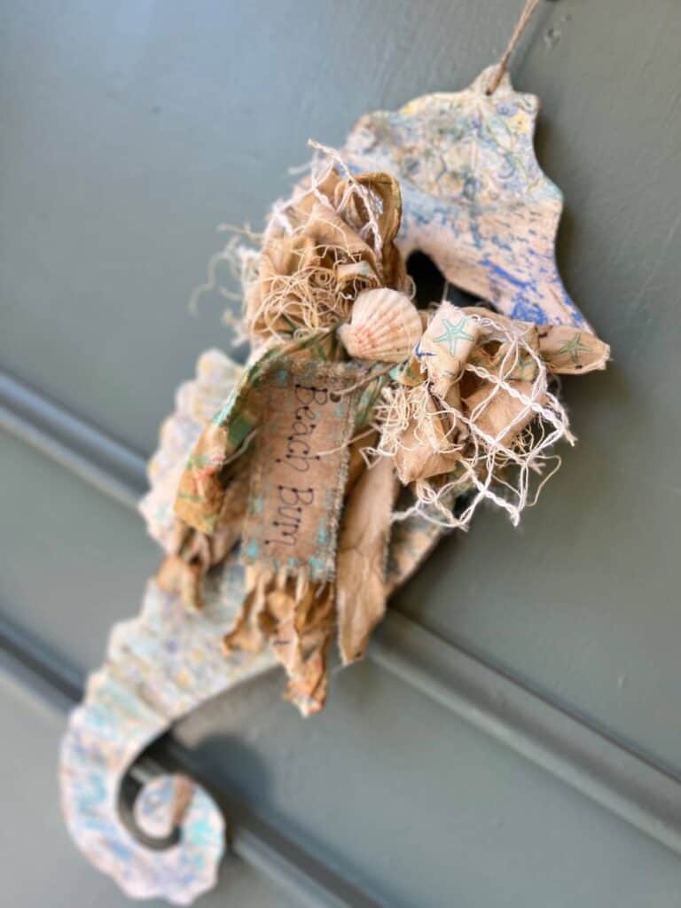Dollar tree chippy seahorse with craftkins makeover of wood seahorse with blue green chipped paint, a seashell napkin decoupaged on it, a big messy fabric bow with a kraft paper tag that says "Beach Bum" hanging on a front door as summer coastal nautical DIY decor.