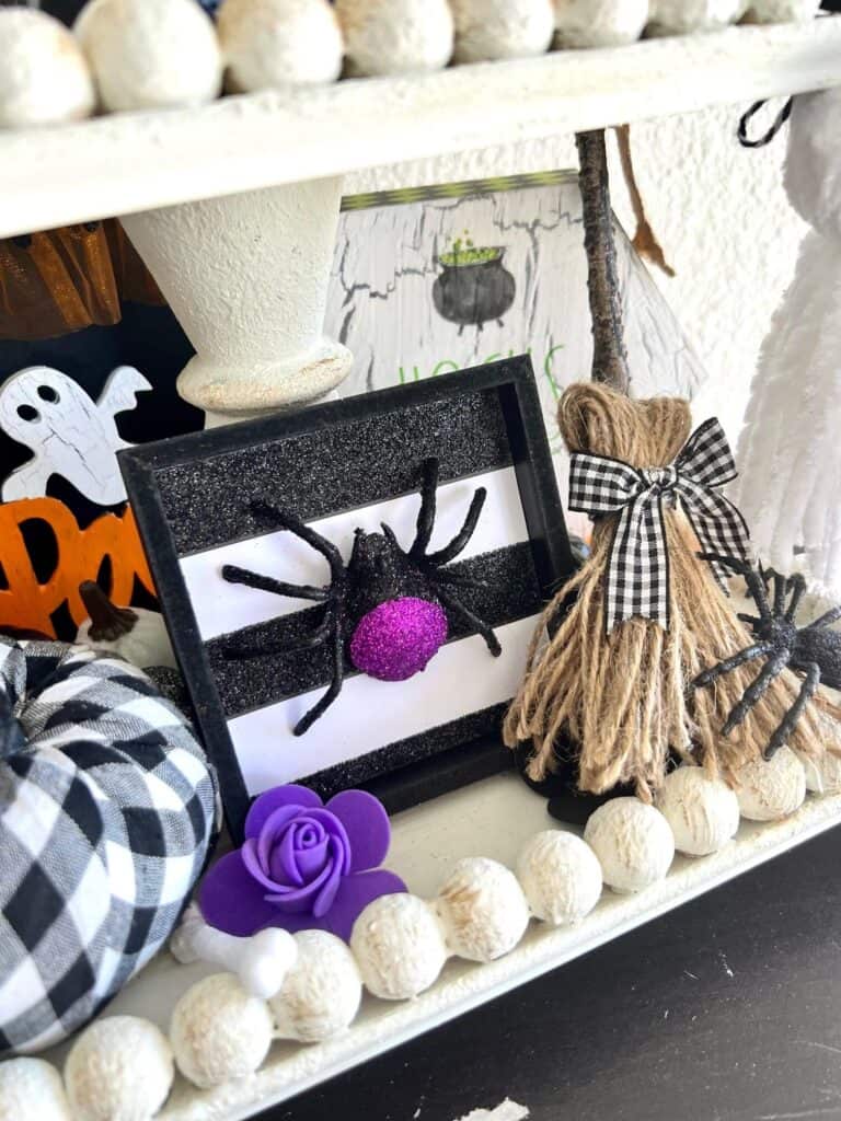 A purple spider on a black and white striped glittery background, next to a twine DIY witches broom.