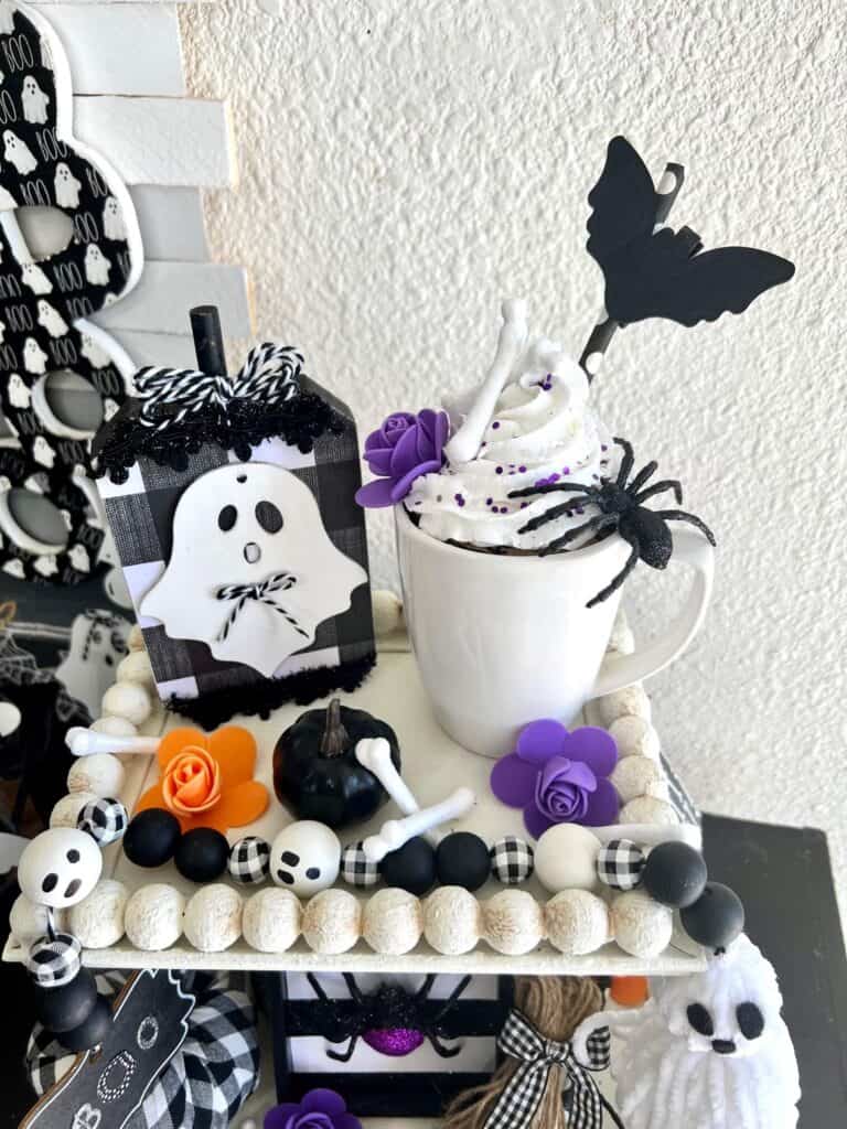 Black and white buffalo check ghost, white faux mug topper with spider, a black pumpkin, and a purple and orange flower on the top of a tiered tray along with a black and white wood bead ghost garland.