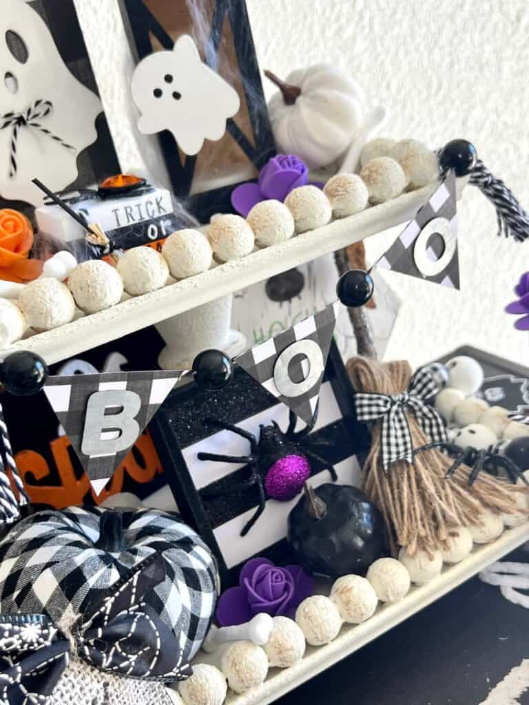 Mini black and white buffalo check BOO banner with tassels on each end, hanging on the top tier of the tray with a purple spider and witch broom underneath.