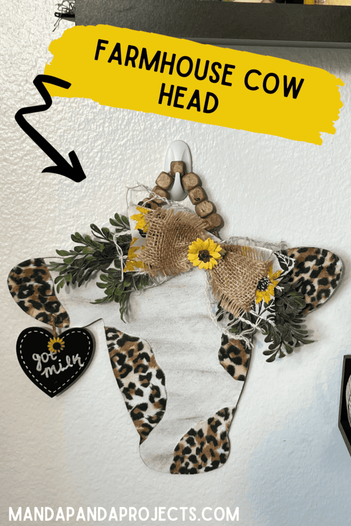 Farmhouse Cow Head with Leopard Print spots, a burlap and sunflower bow with boxwood greenery, a small black heart tag hanging from its ear that says "got milk" and a square wood bead hanger, hanging on the wall as home decor.