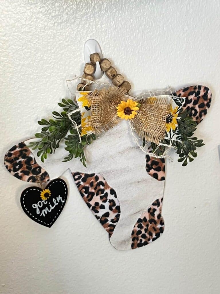 Farmhouse Cow Head with Leopard Print spots, a burlap and sunflower bow with boxwood greenery, a small black heart tag hanging from its ear that says "got milk" and a square wood bead hanger, hanging on the wall as home decor.