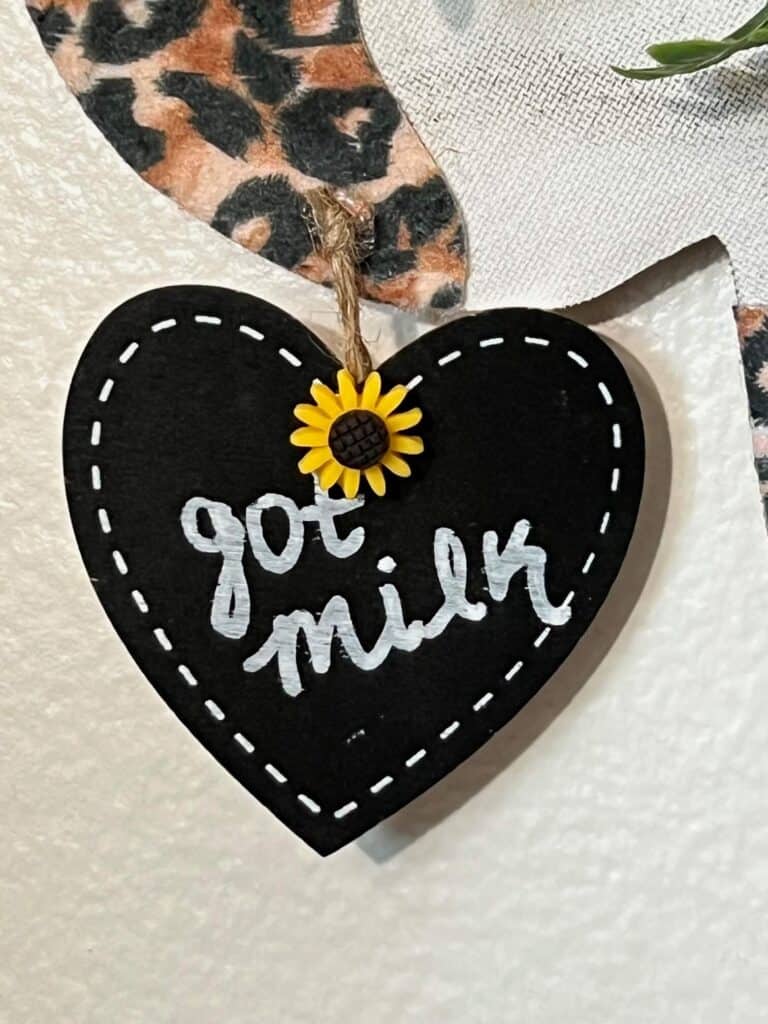 A small black chalkboard heart hangtag that says "got milk" with a small sunflower on the top, hanging from the cows ear with twine.