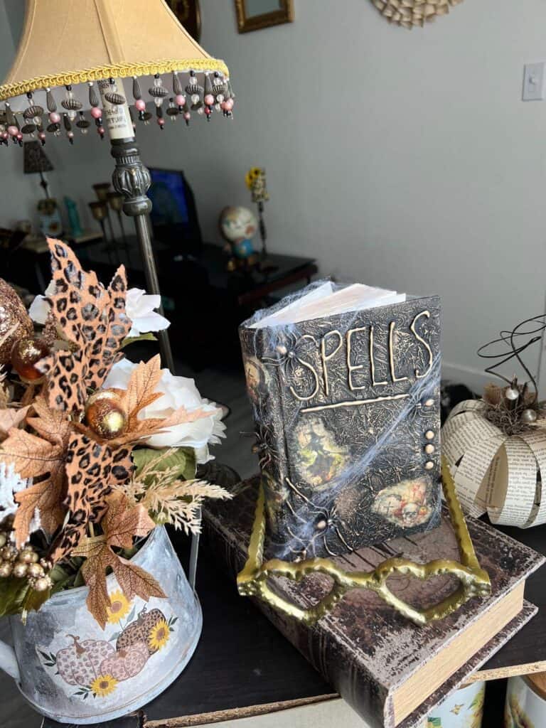The completed and staged spellbook on a bookcase next to a fall floral arrangement and gold heart shaped metal eye glasses.