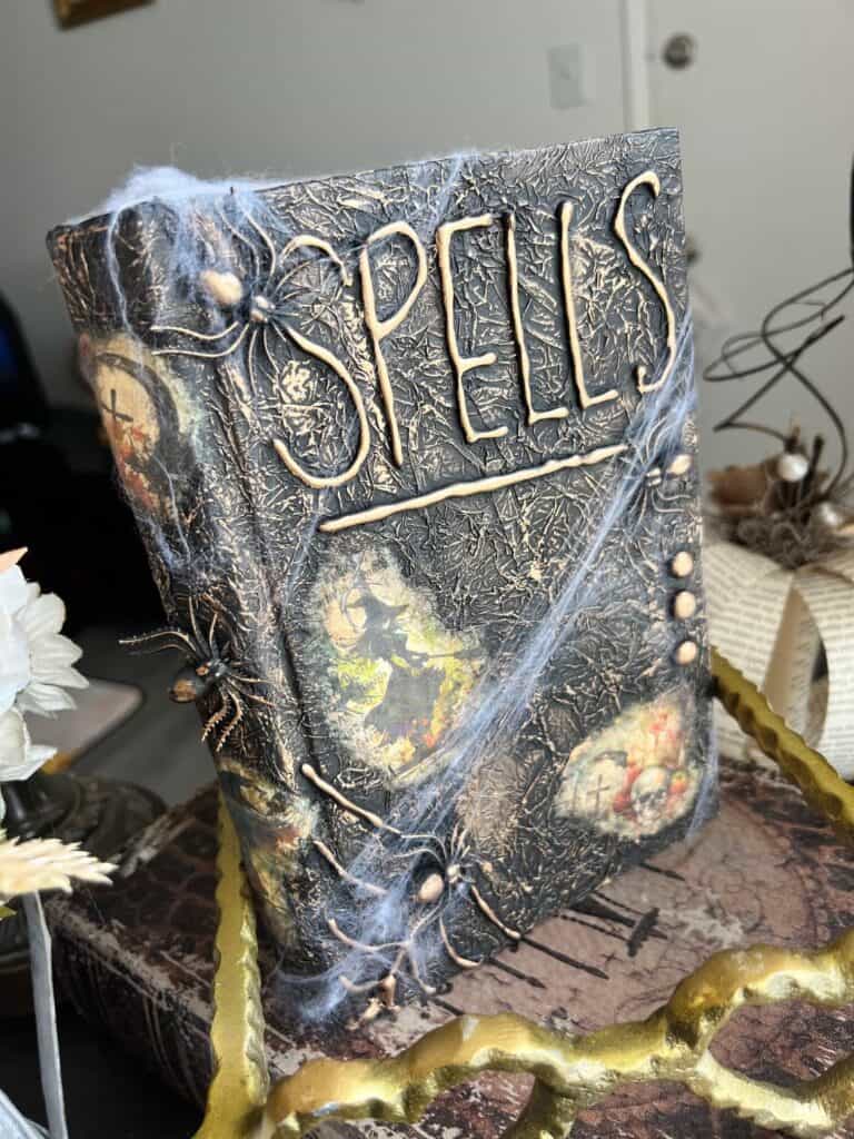 DIY Halloween spellbook decor made with a cheap thrift store book, Digital Deco Designs spell book 1 rice paper with witches, tombstones, and skulls, fake spiders and spiderwebs, all black with antique gold rub and buff on the entire book.