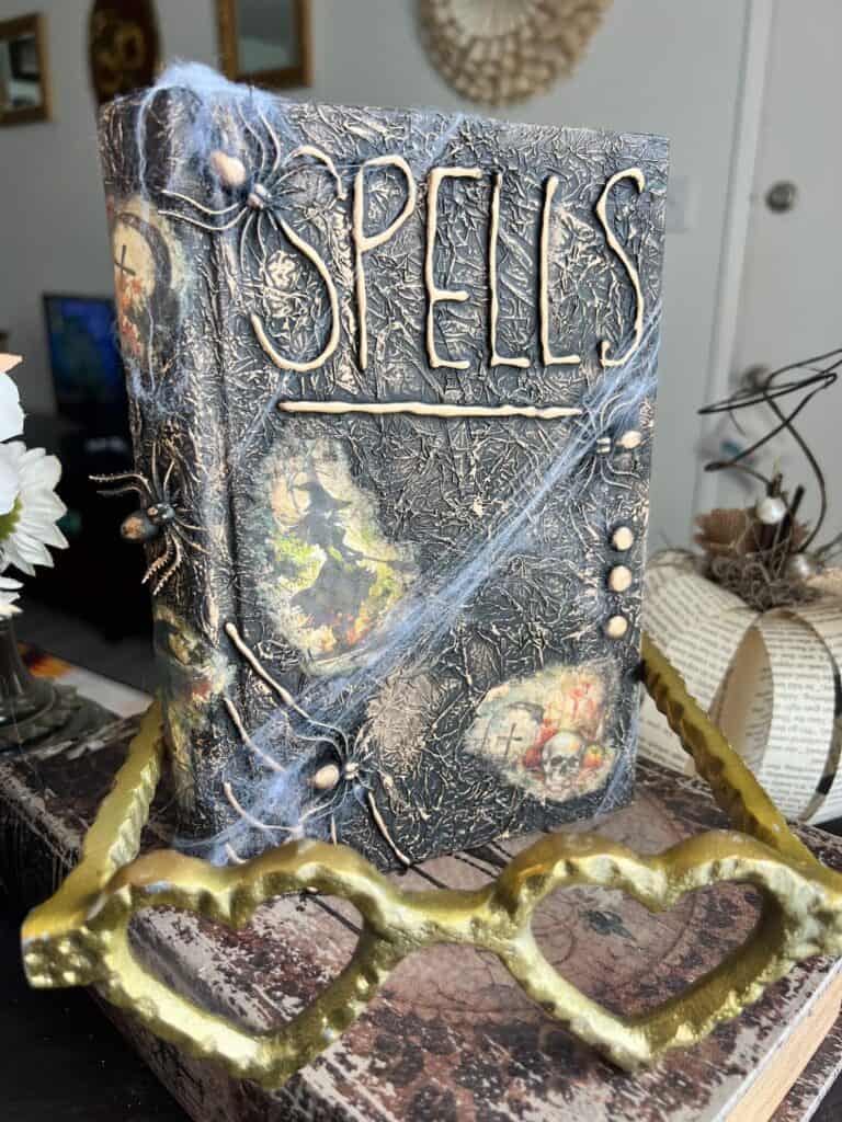 DIY Halloween spellbook decor made with a cheap thrift store book, Digital Deco Designs spell book 1 rice paper with witches, tombstones, and skulls, fake spiders and spiderwebs, all black with antique gold rub and buff on the entire book.