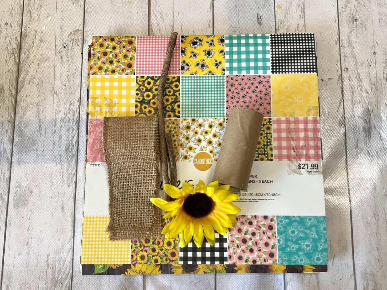 A pink, yellow, teal, and brown sunflower themed scrapbook with burlap, sunflower head, sticks, and a TP roll sitting on a table.