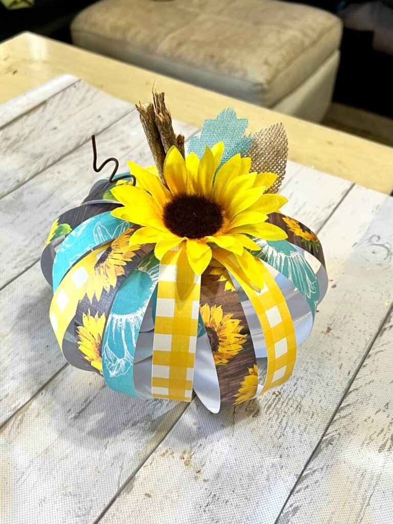 Scrapbook Paper Pumpkin made with a Toilet paper roll, teal, yellow, and brown sunflower scrapbook paper, sticks as a stem, a sunflower head, and a burlap leaf for simple recycled crafts DIY fall decor.