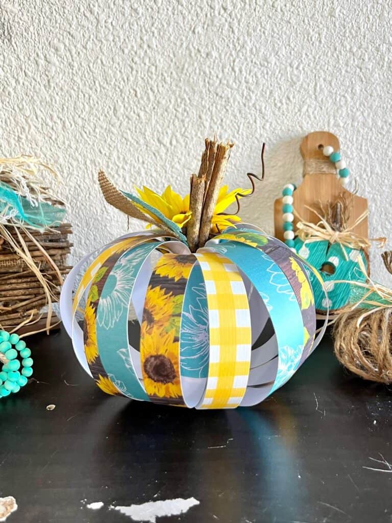 The teal, yellow, and brown sunflower theme scrapbook paper pumpkin on a bookcase surrounded by lots of other DIY pumpkins decor.