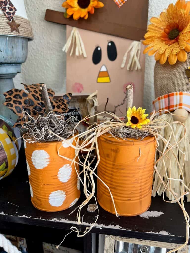 Easy and affordable recycled tin can pumpkin fall decor craft made with just a few cheap supplies a sunflower and a leopard print leaf with a scarecrow and a candy corn nose in the background.