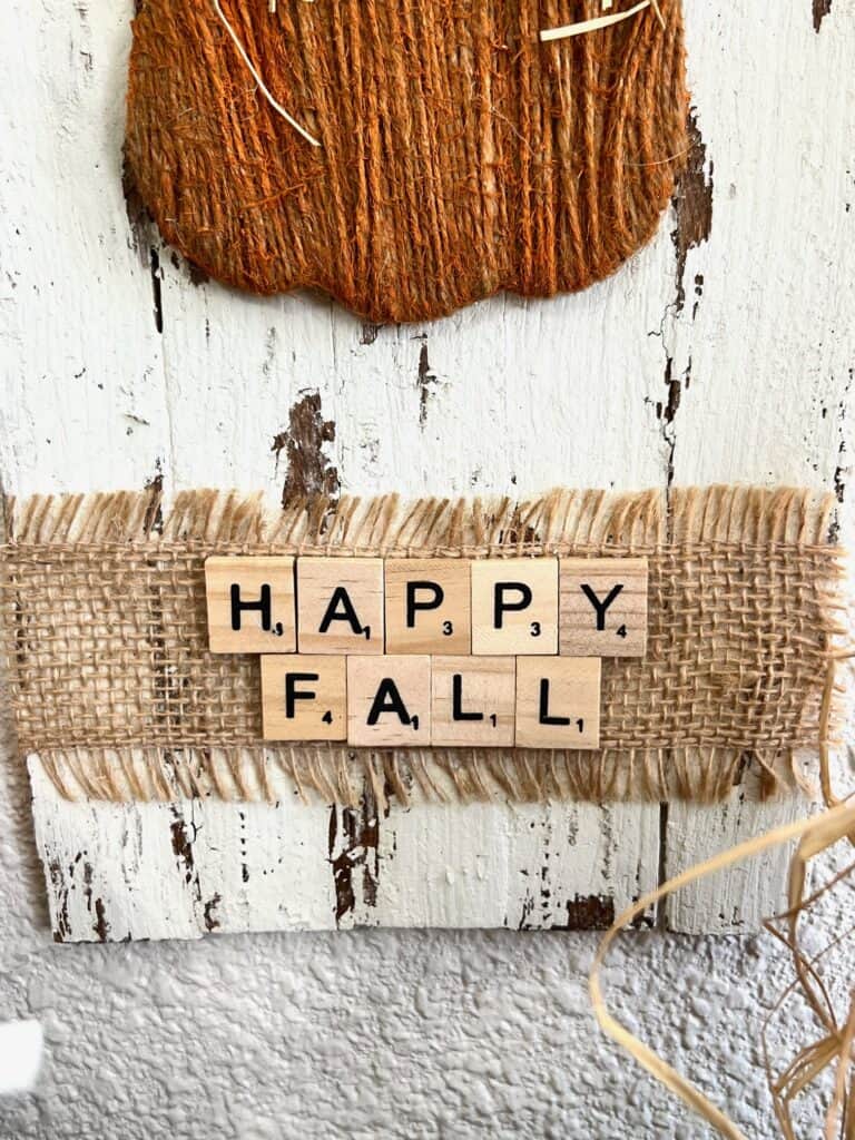 A strip of frayed burlap glued across the bottom of the sign, under the twine pumpkin with scrabble tile letters that say "Happy Fall" glued on top.