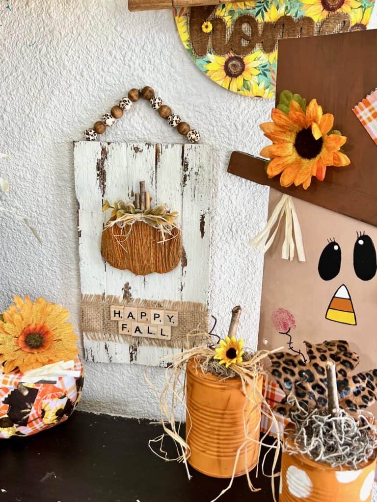 The completed project hanging on the wall with a bookcase underneath that has a patchwork pumpkin, tin can pumpkin, and a wood scarecrow.