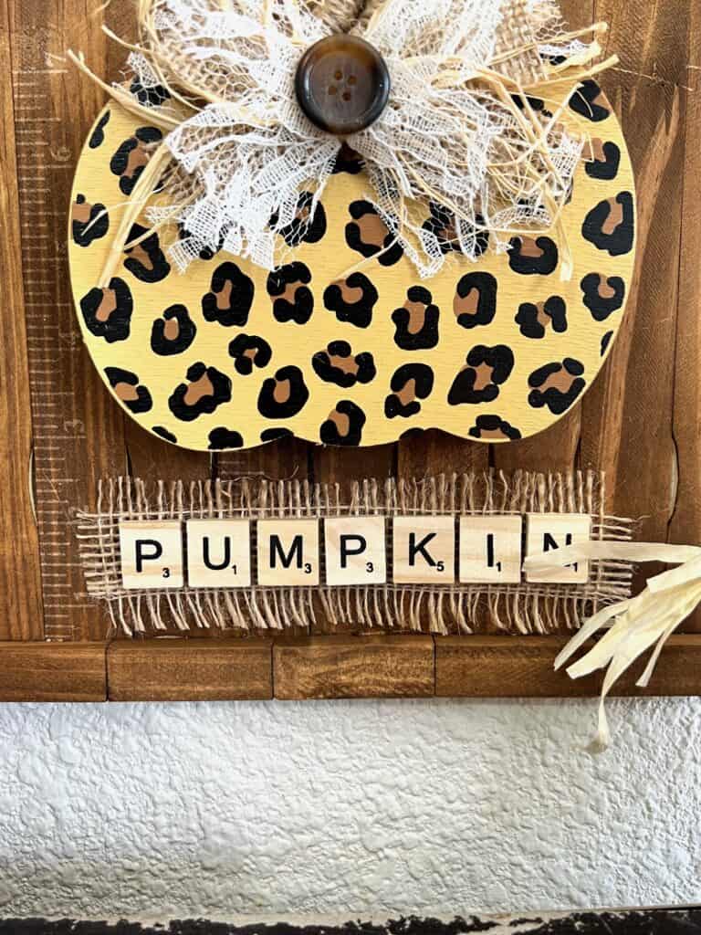A piece of frayed burlap ribbon on the bottom of the sign with the word "PUMPKIN" spelled out in scrabble tiles on top.
