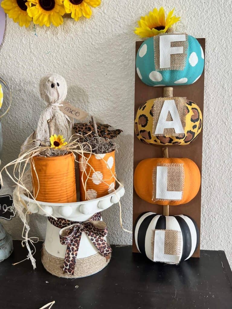 A riser with a primitive ghost and 2 tin can pumpkins sitting on it, with a FALL pumpkin long sign to the right. one teal and white polka dot pumpkin, one leopard print pumpkin, one orange and one black and white striped.