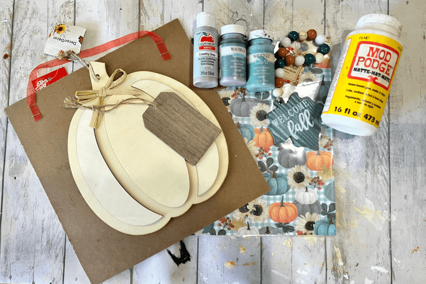 All of the supplies needed for the project laid out on the table. A wood pumpkin, scrapbook paper, wood background, paint, Mod Podge, and wood bead garland.