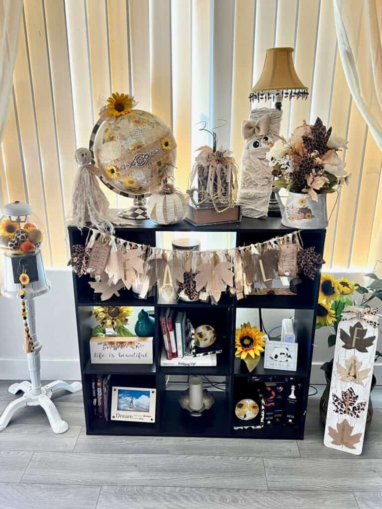 Fall-O-ween decorated book case with a fall banner in the front, a sunflower and leopard print pumpkin filled gumball machine and a Fall sign on the right.