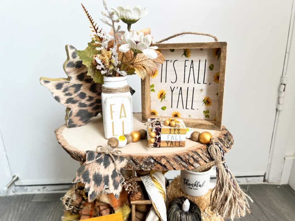 The top tier of a 3 tiered tray with a fall floral salt shaker, a mini book stack, a wood bead garland, and an It's Fall Y'all Sunflower frame.