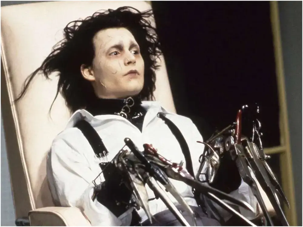 Edward scissorhands laying in a hair dressing chair with his scissorhands showing wearing the gray pants and white shirt with suspenders.