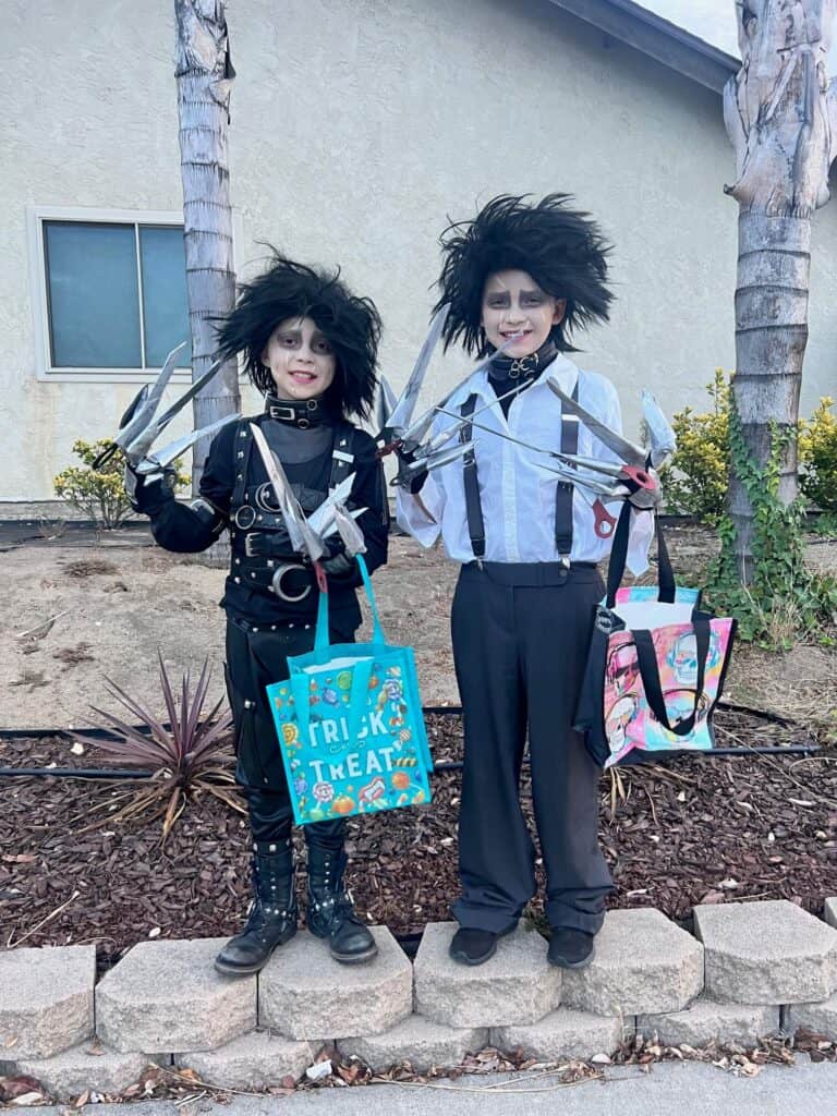 DIY Edward Scissorhands Costumes made with thrift store clothes and belts. Two different versions from the movie, one with black belted leather suit and one with grey pants, and white shirt with suspenders. Both wearing handmade scissorhands gloves and wild black wigs with scars on their faces and a pale complexion. Both kids are holding trick or treat bags.