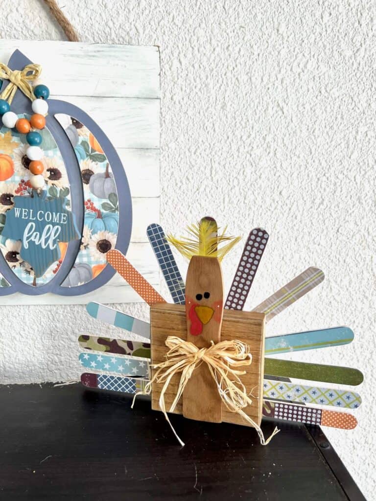 DIY Wood Block Turkey made with a 4x4 wood square, jumbo popsicle sticks, coordinating fall scrapbook paper, a raffia bow, and a cute feather, decor for your thanksgiving holiday.