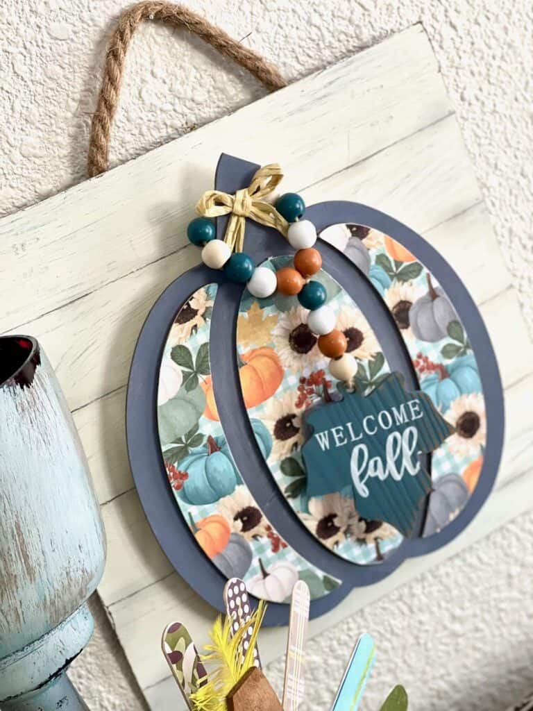 Dollar Tree Fall Wood Pumpkin Sign with a blue grey pumpkin with pretty fall scrapbook paper, a "welcome fall" leaf wood bead garland hanging from the Pumpkin stem, mounted on a faux shiplap background hanging with jute rope.