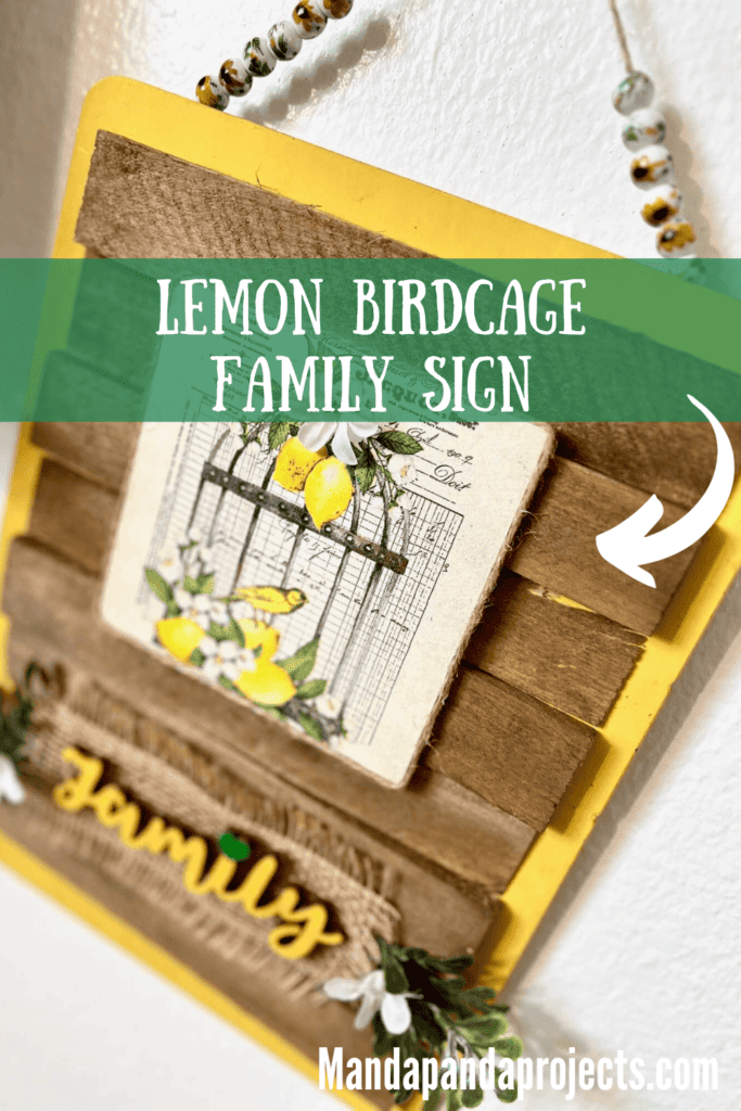 Lemon Birdcage Napkin Family Sign with a yellow background, stained shims, glass sunflower bead hanger, and the word FAMILY painted green at the bottom with greenery and small white flowers for hanging DIY home decor.