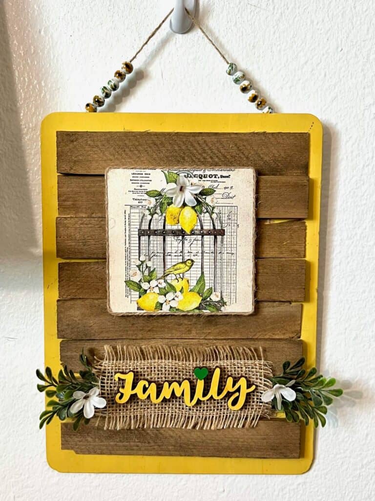 Lemon Birdcage Napkin Family Sign with a yellow background, stained shims, glass sunflower bead hanger, and the word FAMILY painted green at the bottom with greenery and small white flowers for hanging DIY home decor.