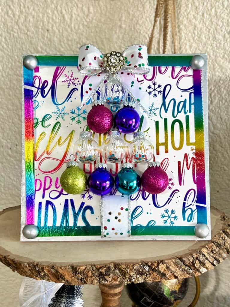 Colorful Ornament bulb Christmas Tree Frame made with a holiday rainbow color napkin with various christmas phrases on it, small round bulb ornaments in the shape of a christmas tree with a bow as the tree topper and som totally dazzled bling. Bright and cheery diy christmas decor.