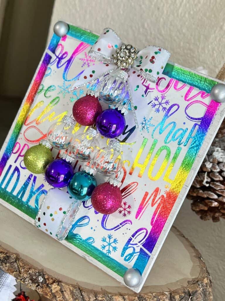 Colorful Ornament bulb Christmas Tree Frame made with a holiday rainbow color napkin with various christmas phrases on it, small round bulb ornaments in the shape of a christmas tree with a bow as the tree topper and som totally dazzled bling. Bright and cheery diy christmas decor.