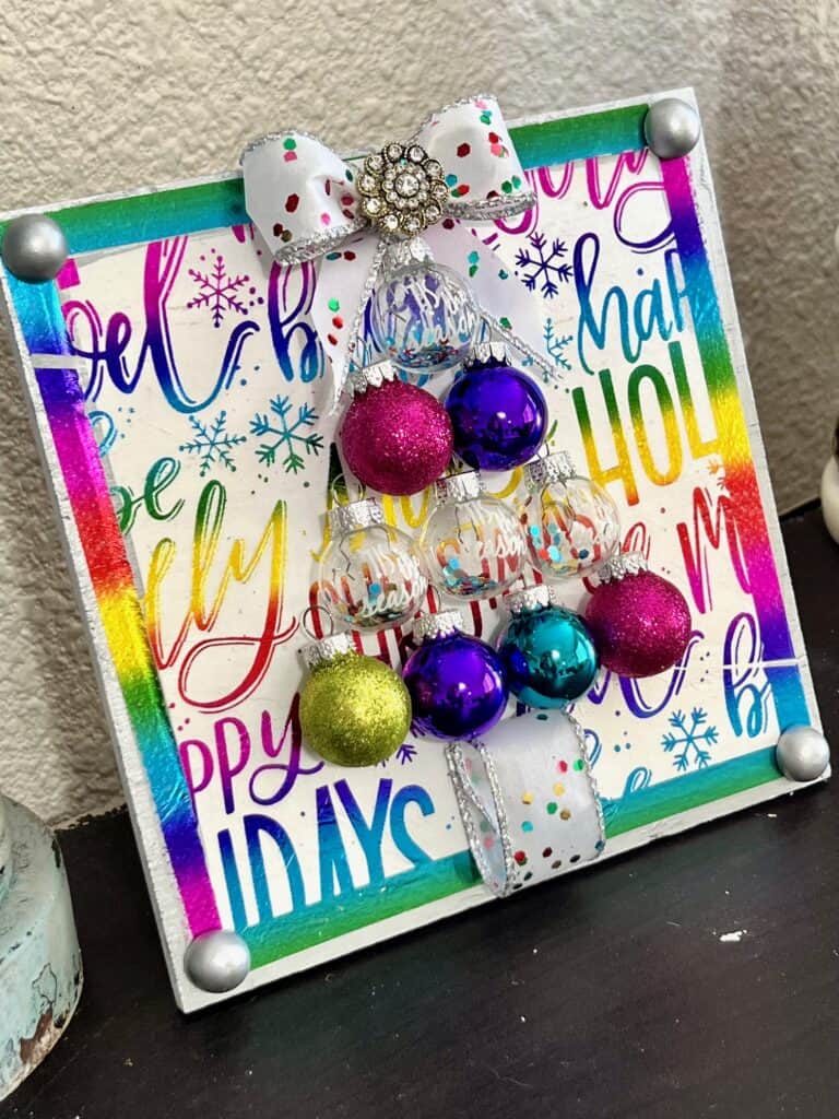 Colorful Ornament bulb Christmas Tree Frame made with a holiday rainbow color napkin with various christmas phrases on it, small round bulb ornaments in the shape of a christmas tree with a bow as the tree topper and som totally dazzled bling. Bright and cheery diy christmas decor.