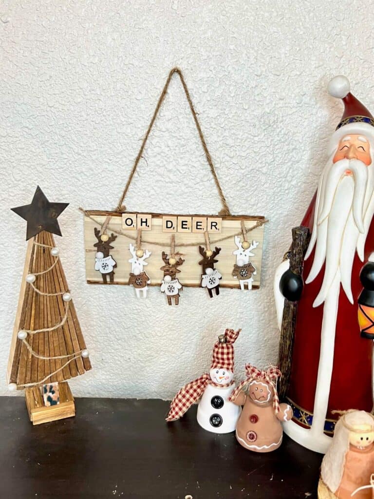 The OH Deer oversized ornament hanging on the wall next to a wooden shim christmas tree with pearls and a star, next to a Santa, gingerbread man, and snowman on a bookcase.