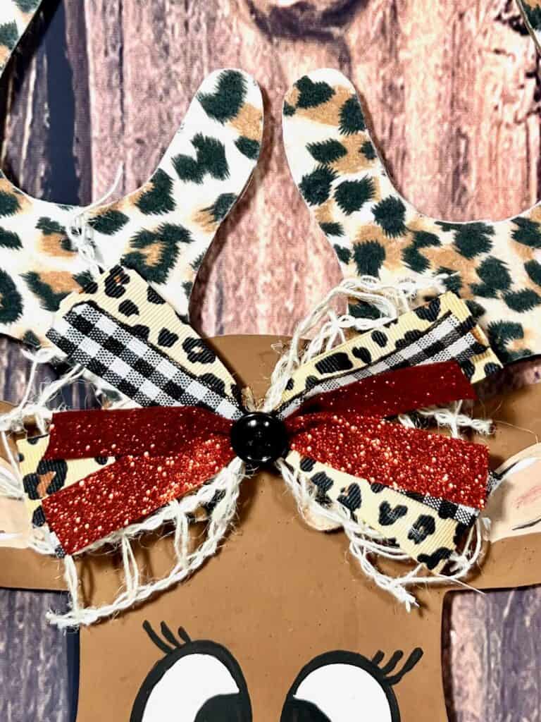 Cute bow with red glitter ribbon, buffalo check, and leopard print, in between the reindeers ears and leopard print antlers.