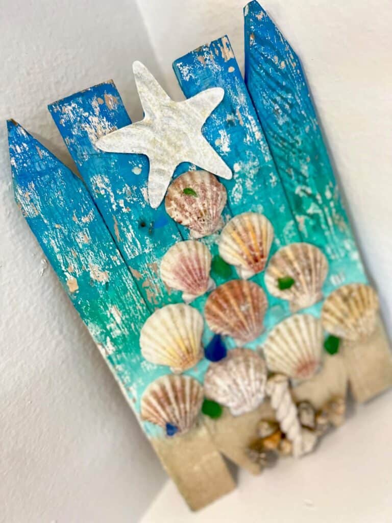 Seashell Coastal Christmas Tree with blue and green Ombre background, real sand and shells at the bottom, seashells in the shape of a Christmas tree, and a white sparkly starfish on top, with a nautical rope tree trunk. DIY coastal nautical Christmas in July decor.