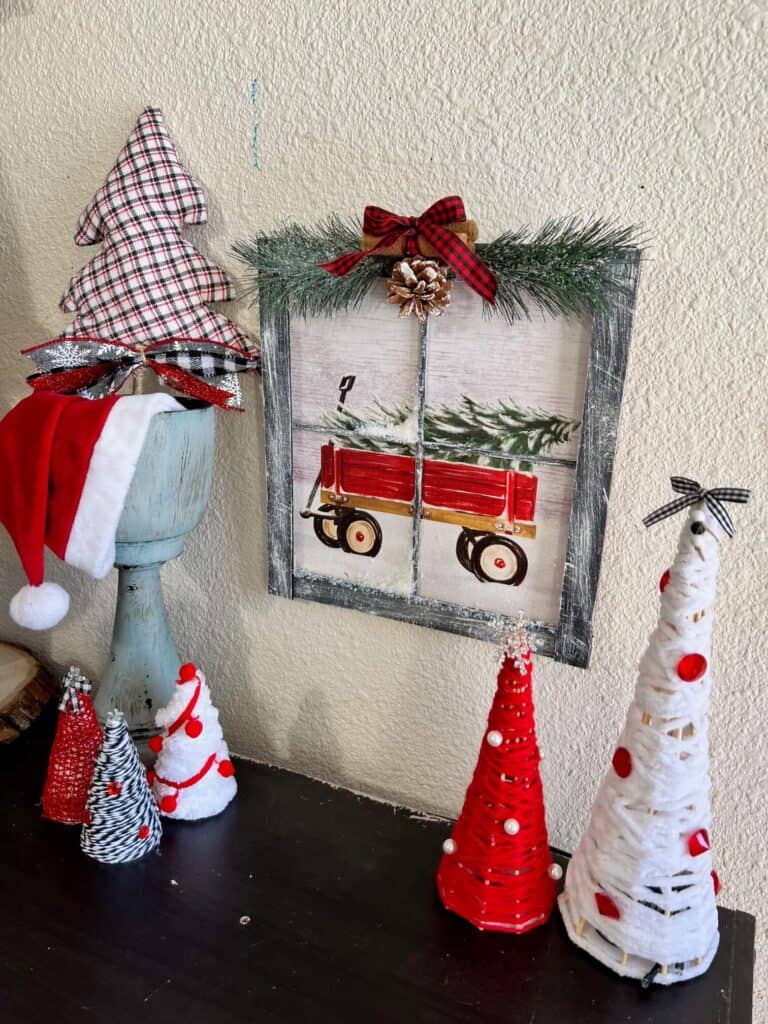 The christmas wagon hanging on the wall with foam christmas trees to the left and tall red and white yarn christmas trees to the right on a bookshelf.