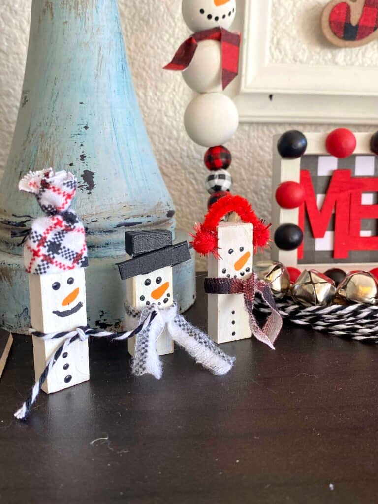 Jenga Block Snowmen made with Dollar Tree tumbling tower blocks, painted white with simple hand drawn faces, and decorated with fabric and twine scarves, hats, top hats, and ear muffs to display on your Christmas and winter themed DIY tiered trays. 