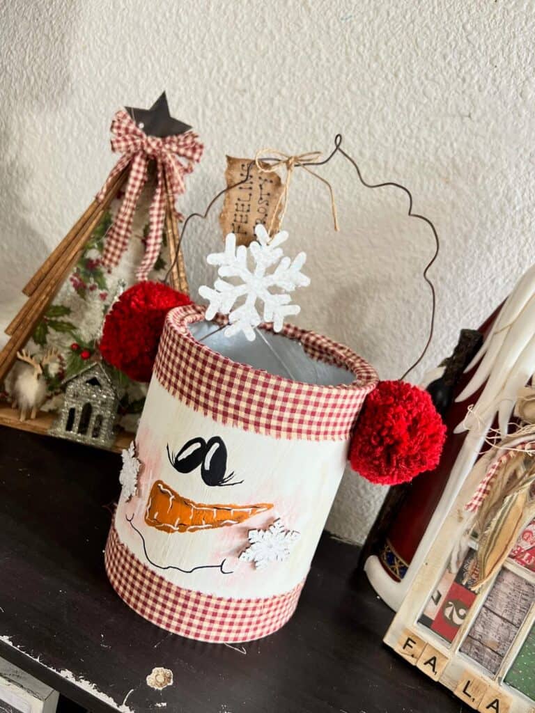 Coffee can snowman made with a recycled coffee can, rusty wire, red pom pom ear muffs, a hand painted face, snowflakes and a hangtag that says "Feeling frosty" for DIY christmas and winter crafts and decor.