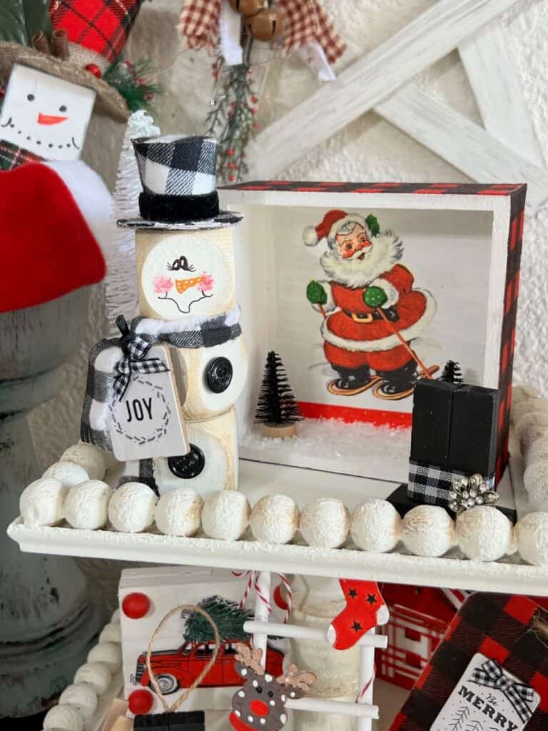 The wood block snowman on the top tier of a tiered tray next to a retro santa frame.