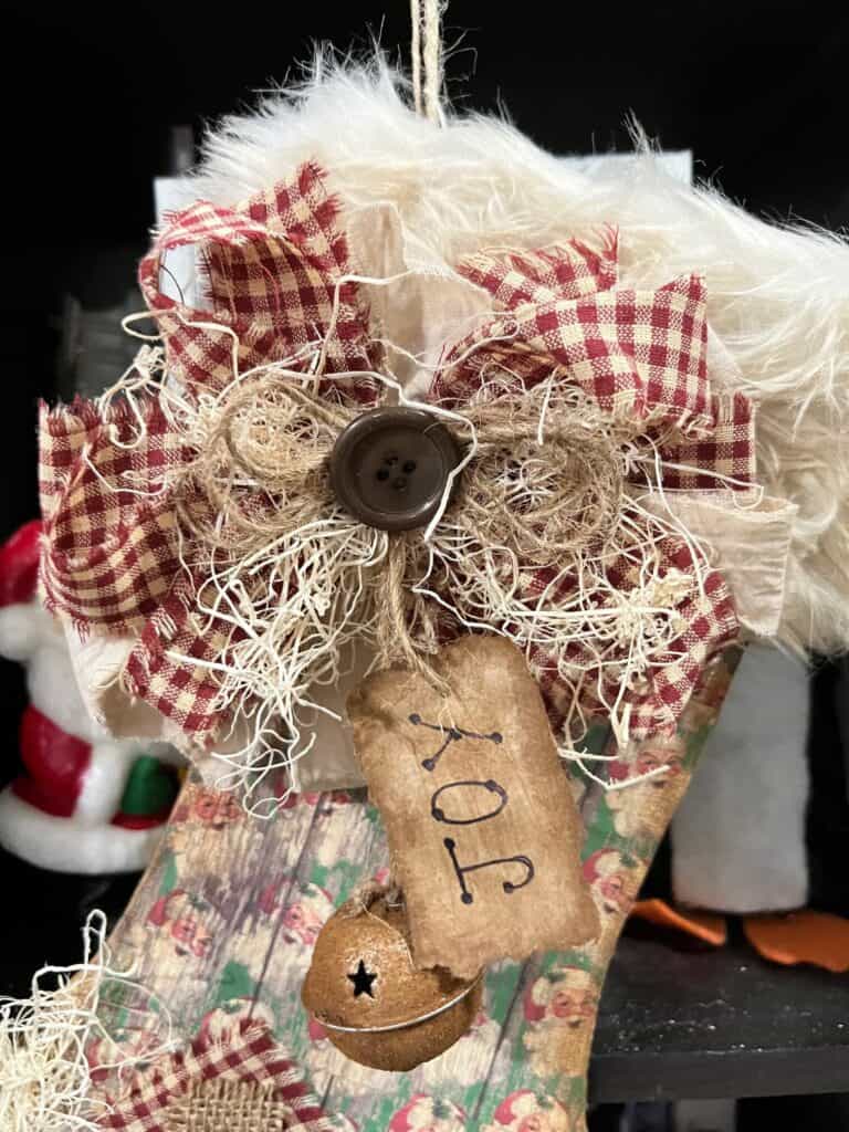 A big messy fabric bow with a hangtag that says "Joy" made from kraft paper and a rusty bell hanging from the bow on the top of the stocking.