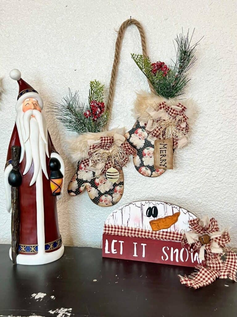 A tall vintage santa on the left, the Hanging Christmas Mittens in the middle hanging on the wall, and a dollar tree snowman on the right on a bookshelf.