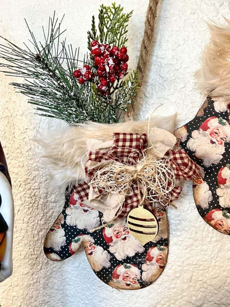 Close up of the DIY christmas mittens of the mitten on the right with the greenery and berries coming out the top of the fur cuff and a small bow with a small hanging gold ornament.