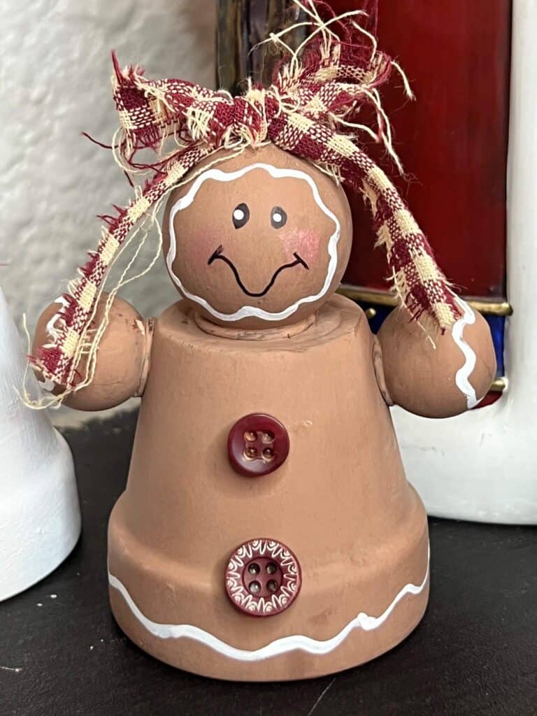 Mini terra cotta pot gingerbread man painted brown with a re homespun fabric bow on top of its bead head, red buttons and a simple hand painted face.