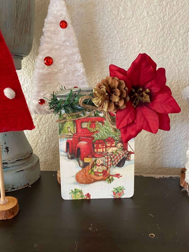 Christmas Napkin Wood Mason Jar with a napkin with the red truck with presents in the bed, some faux pine on the top with a gold glitter pine cone and a big poinsettia.