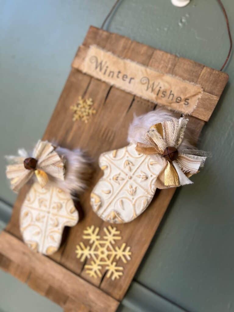 Wooden Winter Mittens Decor on a stained paint stick background, white and gold mittens with grungy faux fur, a small bow on the cuff of each, gold snowflakes, and the top says "Winter Wishes".