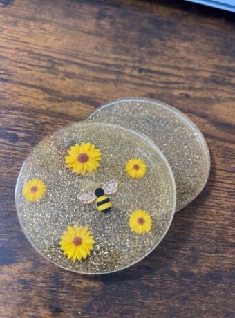 2 resin coaster, one with gold glitter and sunflowers and a bee and the other one with just gold glitter sitting on a table overlapping one another.