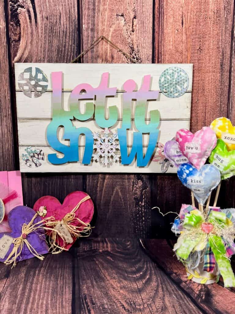 The Let it Snow sign hanging on a wood backdrop with pink and purple hearts to the left and a pastel rainbow bouquet of stuffed hearts to the right.